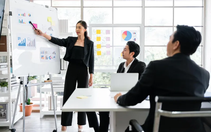 An analyst presenting charts at a whiteboard to two seated colleagues